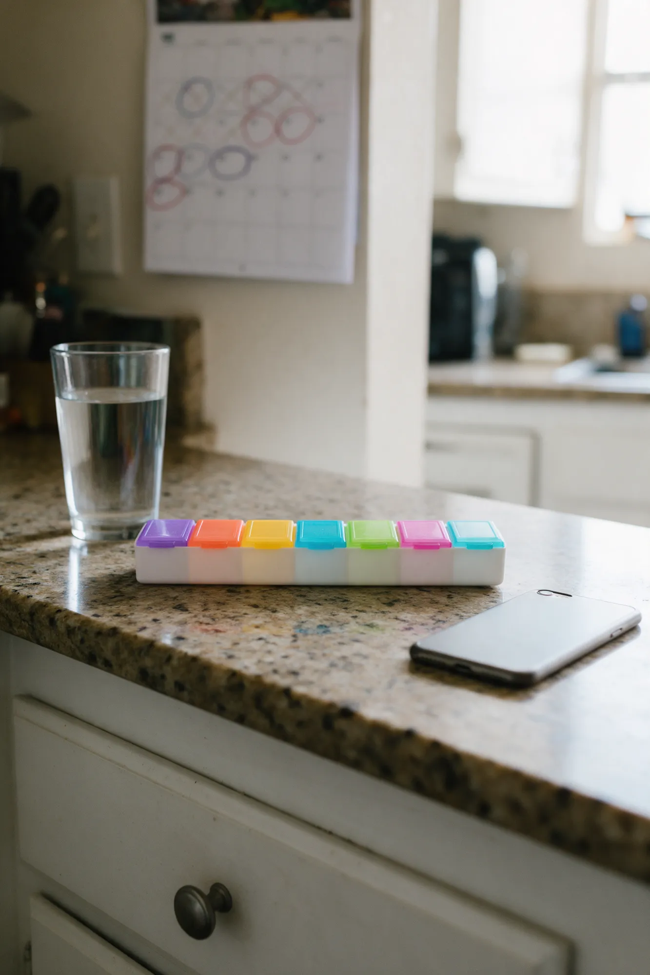 A pill organizer, a glass of water and a phone on a kitchen counter, with a calendar of circled appointments behind