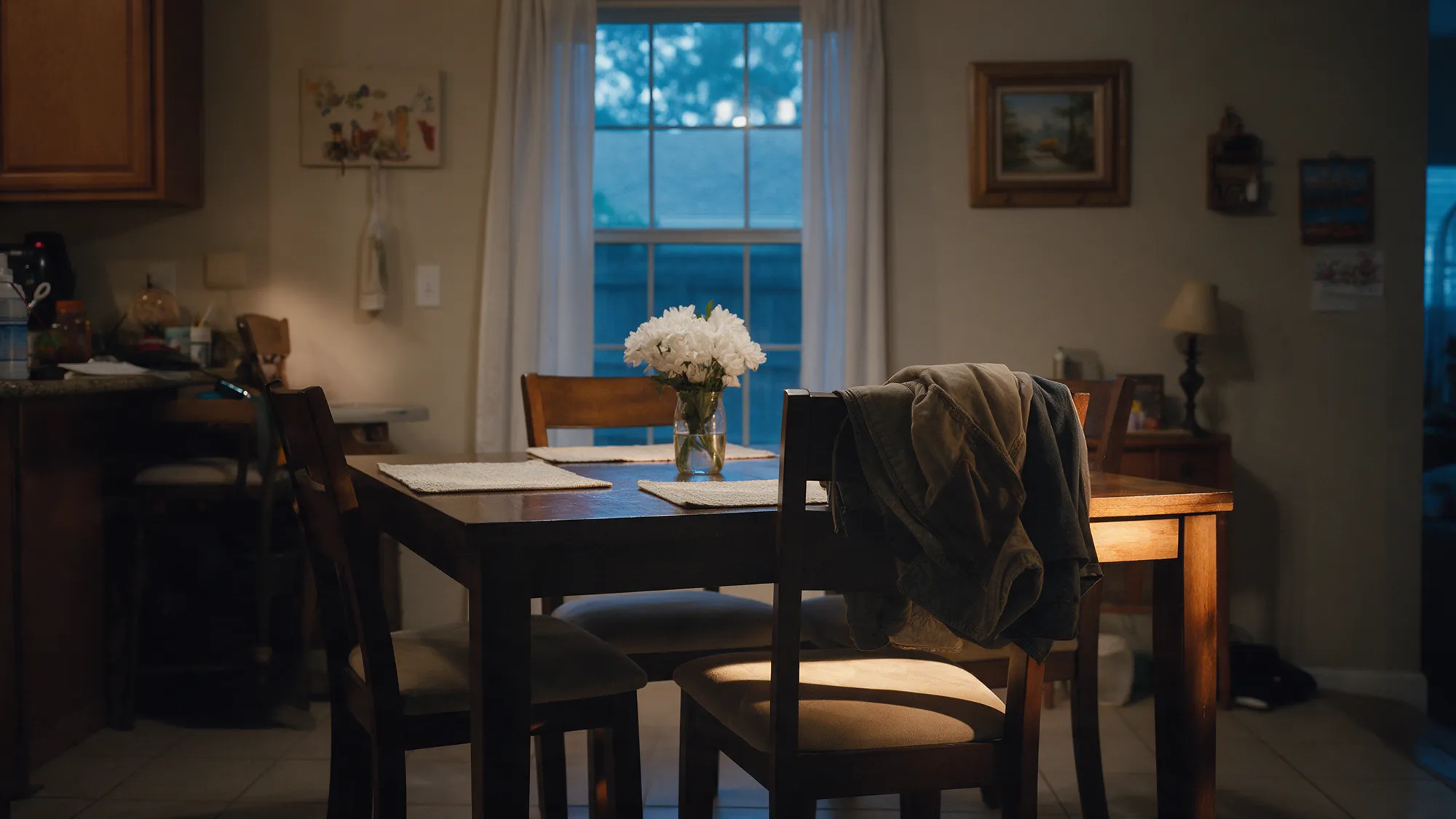A family dining table at dusk with one empty chair and a vase of white flowers